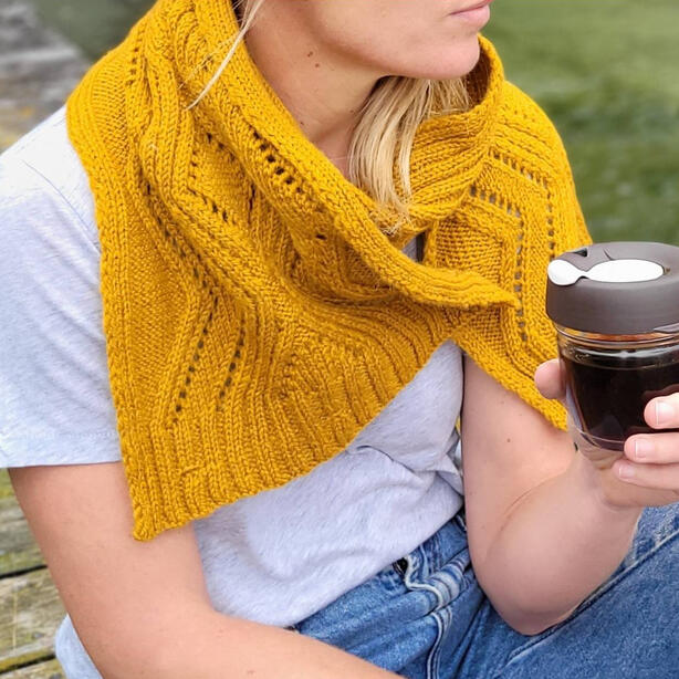 Women wearing a deep gold or mustard coloured wide scarf with lacework and cable details and holding a coffee sitting on a wooden pier