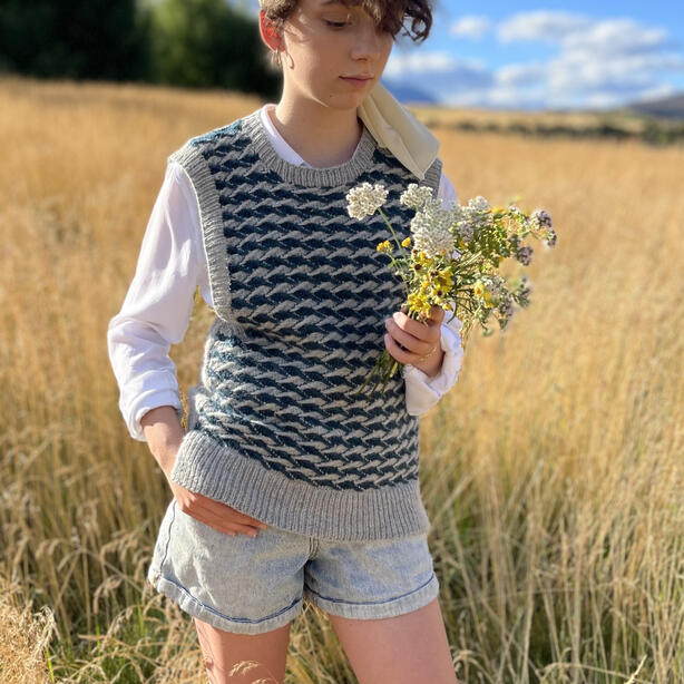 Young woman standing in a field holding flowers and wearing a textured hand knitted vest of jacquard stitch